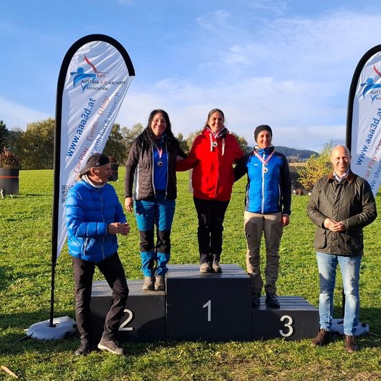 Fünf Personen stehen auf einem Podest auf einem Außenfeld. Sie lächeln und tragen Medaillen. Zwei Banner stehen hinter ihnen. Sie posieren wahrscheinlich für ein Foto.