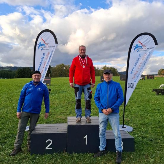 Drei Männer stehen auf einem Podium, einer in einem roten Jacken mit einer Medaille, und die anderen in blauen Jacken. Sie sind draußen auf einem Rasenfeld mit Bannern hinter ihnen.