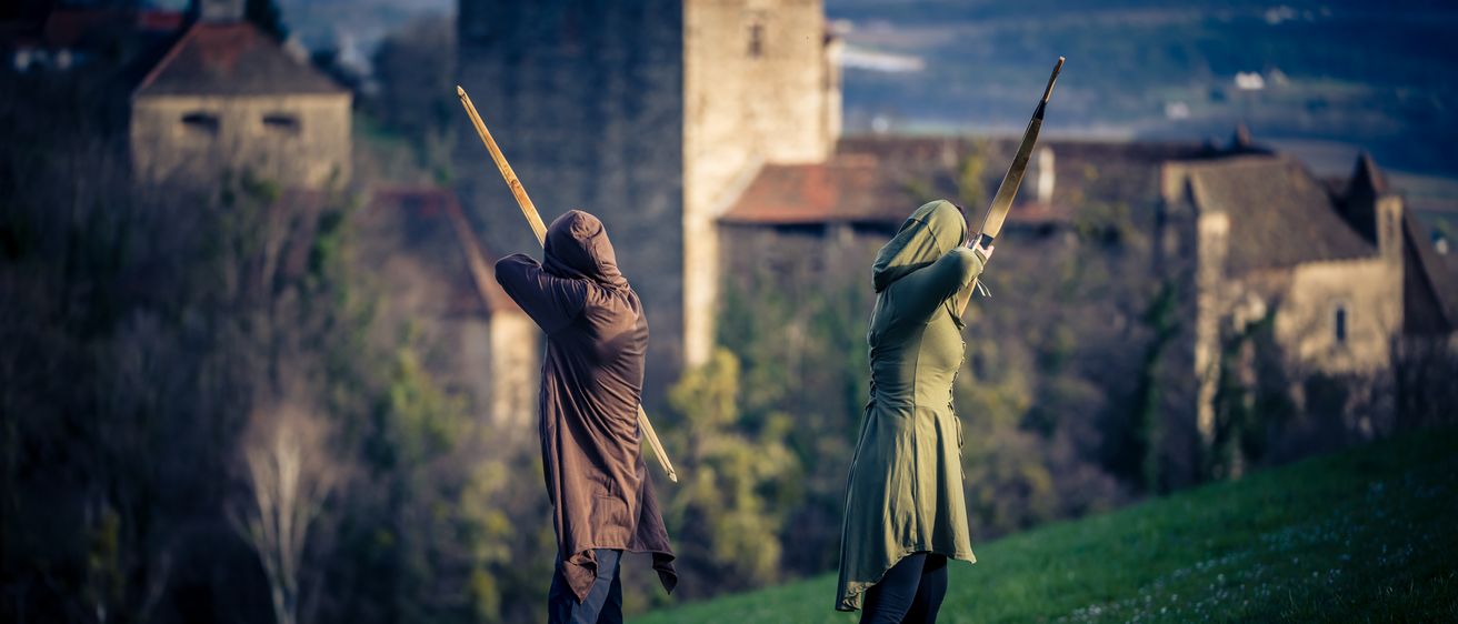 Zwei Personen stehen auf einem Hügel und halten jeweils einen Bogen und Pfeil in der Hand, während sie eine Burg in der Ferne überblicken.