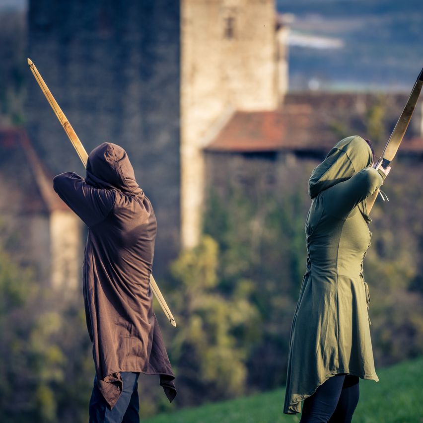 Zwei Personen stehen auf einem Hügel und halten jeweils einen Bogen und Pfeil in der Hand, während sie eine Burg in der Ferne überblicken.