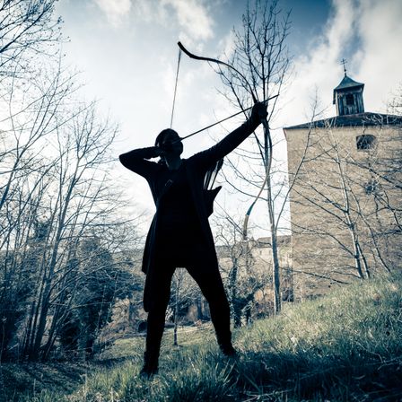 Eine silhouettierte Figur, gekleidet in dunkler Kleidung, hält einen Bogen mit gespannter Sehne und zielt auf den Himmel. Sie steht auf einem Grashügel mit kahlen Bäumen und einer Burg im Hintergrund.