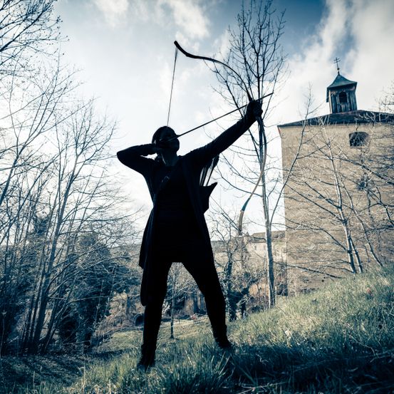 Eine silhouettierte Figur, gekleidet in dunkler Kleidung, hält einen Bogen mit gespannter Sehne und zielt auf den Himmel. Sie steht auf einem Grashügel mit kahlen Bäumen und einer Burg im Hintergrund.