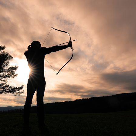 Die Silhouette einer Person, die einen Bogen und einen Pfeil hält, zielt auf die Sonne in einem Feld bei Sonnenuntergang.