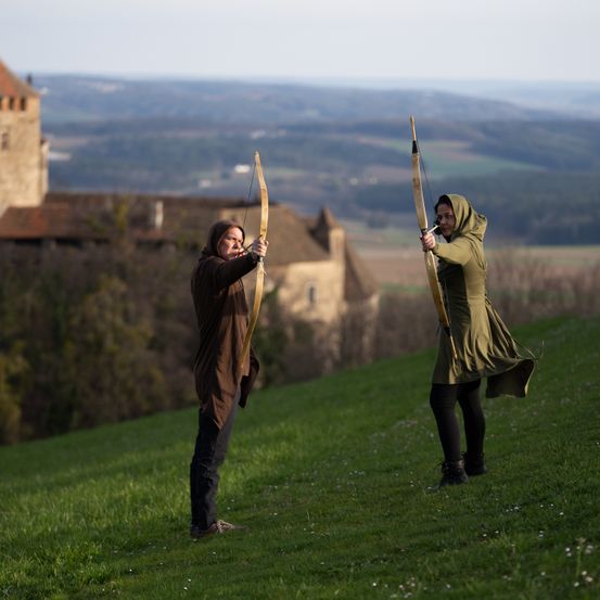 Zwei Personen stehen auf einem Grashügel, jede mit einem Bogen in der Hand, mit einer Burg im Hintergrund unter einem klaren Himmel.