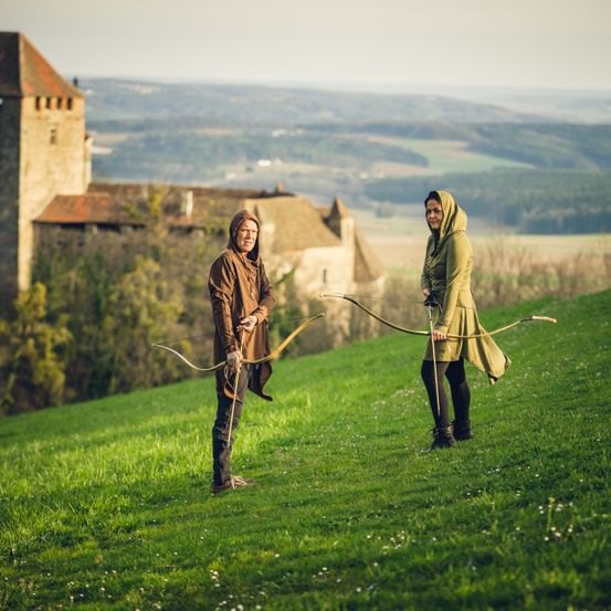 Zwei Personen in mittelalterlicher Kleidung stehen auf einem Grashügel und halten Bögen. Hinter ihnen ist ein Schloss mit einem Turm zu sehen, vor einem Hintergrund von Hügeln und klarem Himmel.