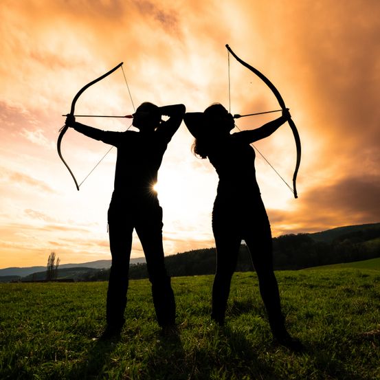 Zwei silhouettierte Bogenschützen, die einander gegenüberstehen, halten ihre Bögen in der Dämmerung, die Sonne geht hinter ihnen unter.