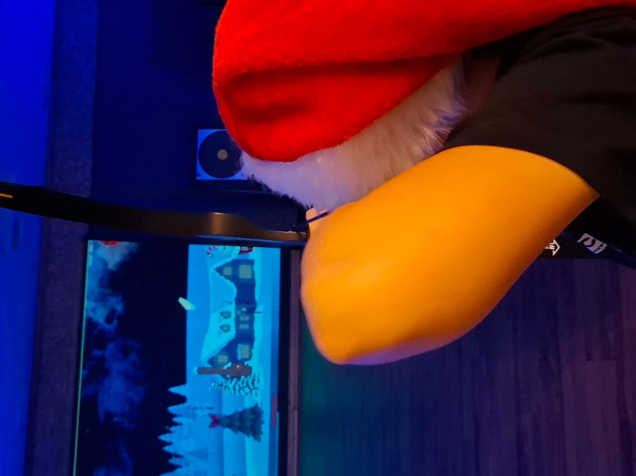 Ein Nahaufnahme eines Weihnachtsmann-Kostüms mit rotem Hut und gelbem Arm, von der Seite betrachtet. Ein blauer Bildschirm zeigt eine Winterszene mit einem Haus und Bäumen.