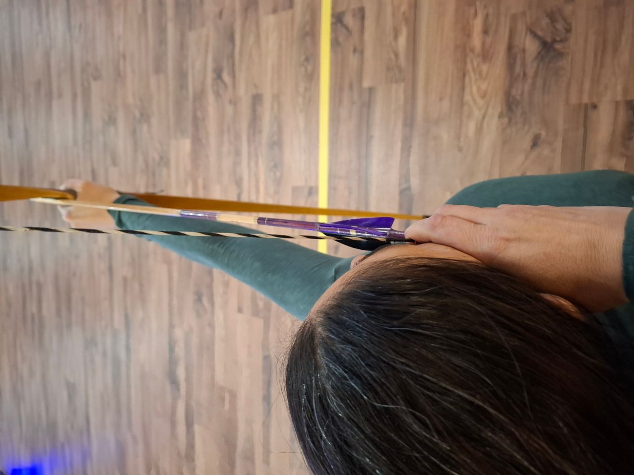 Eine Person hält einen Bogen mit beiden Händen, zielt nach unten und ist in grüner Kleidung gekleidet. Der Boden ist aus Holz, und eine gelbe Linie befindet sich an der Wand.