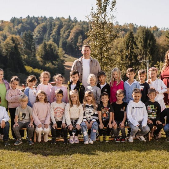 Eine Gruppe von Kindern und Lehrern posiert für ein Foto in einem Grasbereich mit einem Baum und Bergen im Hintergrund.