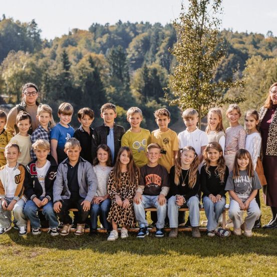 Eine Gruppe von Kindern und zwei Lehrern posiert für ein Foto im Park. Die Kinder sitzen auf einer Bank und lächeln. Bäume sind im Hintergrund.