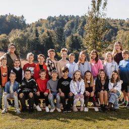 Eine Gruppe von Kindern und Erwachsenen posiert für ein Foto in einem Grasbereich mit einer Baumkulisse. Die Kinder sitzen auf einer Bank, und einige Erwachsene stehen dahinter.