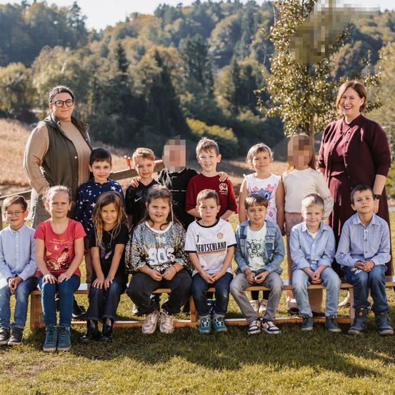 Eine Gruppe von Kindern und zwei Erwachsenen, wahrscheinlich Lehrer, sitzen auf einer Bank in einem Grasfeld. Die Kinder tragen Freizeitkleidung und die Erwachsenen lächeln. Bäume und ein Hügel sind im Hintergrund zu sehen.