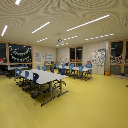 Ein leerer Klassenraum mit gelbem Boden und weißen Tischen. Der Raum ist gut beleuchtet mit Leuchtstoffröhren an der Decke. Es gibt Glasfenster mit Dekorationen.