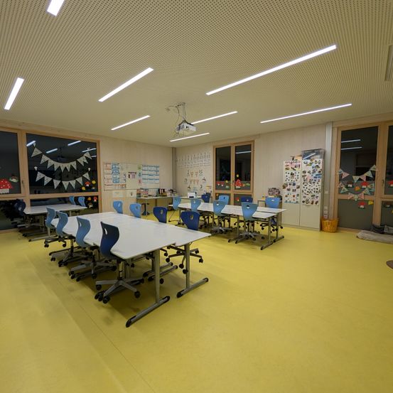 Ein leerer Klassenraum mit gelbem Boden und weißen Tischen. Der Raum ist gut beleuchtet mit Leuchtstoffröhren an der Decke. Es gibt Glasfenster mit Dekorationen.