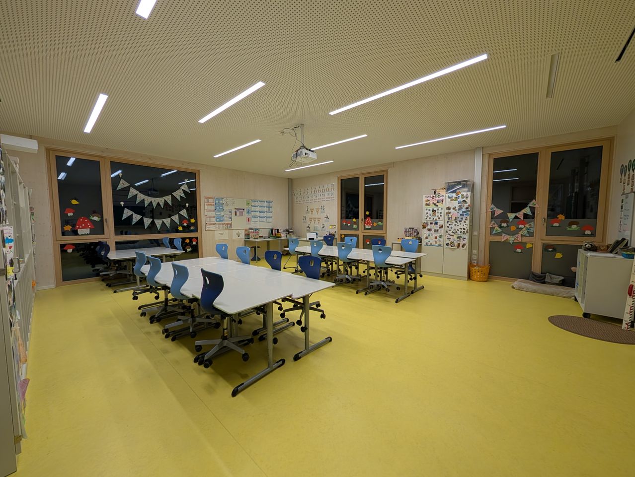 Ein leerer Klassenraum mit gelbem Boden und weißen Tischen. Der Raum ist gut beleuchtet mit Leuchtstoffröhren an der Decke. Es gibt Glasfenster mit Dekorationen.