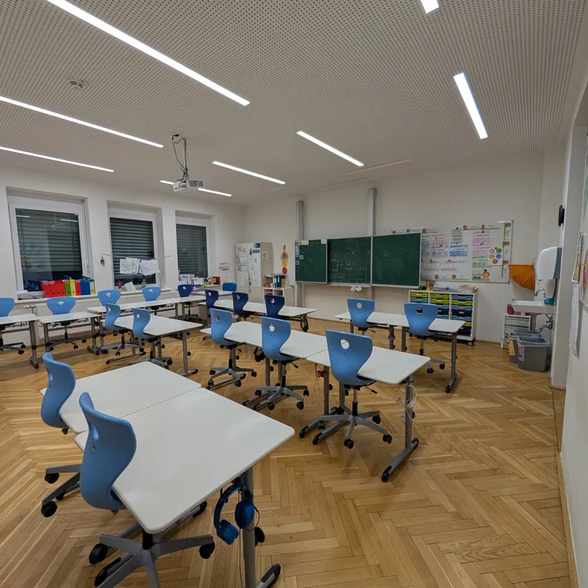 Ein leerer Klassenraum mit weißen Tischen und blauen Stühlen. An der Wand befinden sich drei Tafeln und eine benachbarte Tafel. Fenster sind mit Jalousien verdeckt.