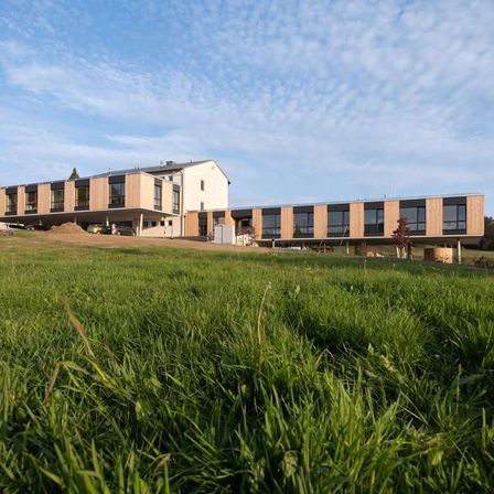 Ein modernes Gebäude mit großen Fenstern und Holzakzenten befindet sich auf einem grünen Feld unter einem teilweise bewölkten Himmel.