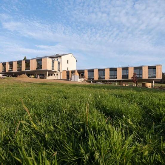 Ein modernes Gebäude mit großen Fenstern und Holzakzenten befindet sich auf einem grünen Feld unter einem teilweise bewölkten Himmel.