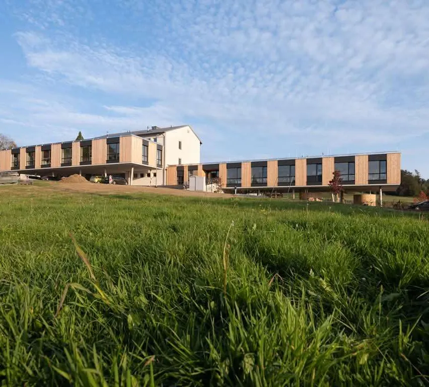 Ein modernes Gebäude mit großen Fenstern und Holzakzenten befindet sich auf einem grünen Feld unter einem teilweise bewölkten Himmel.