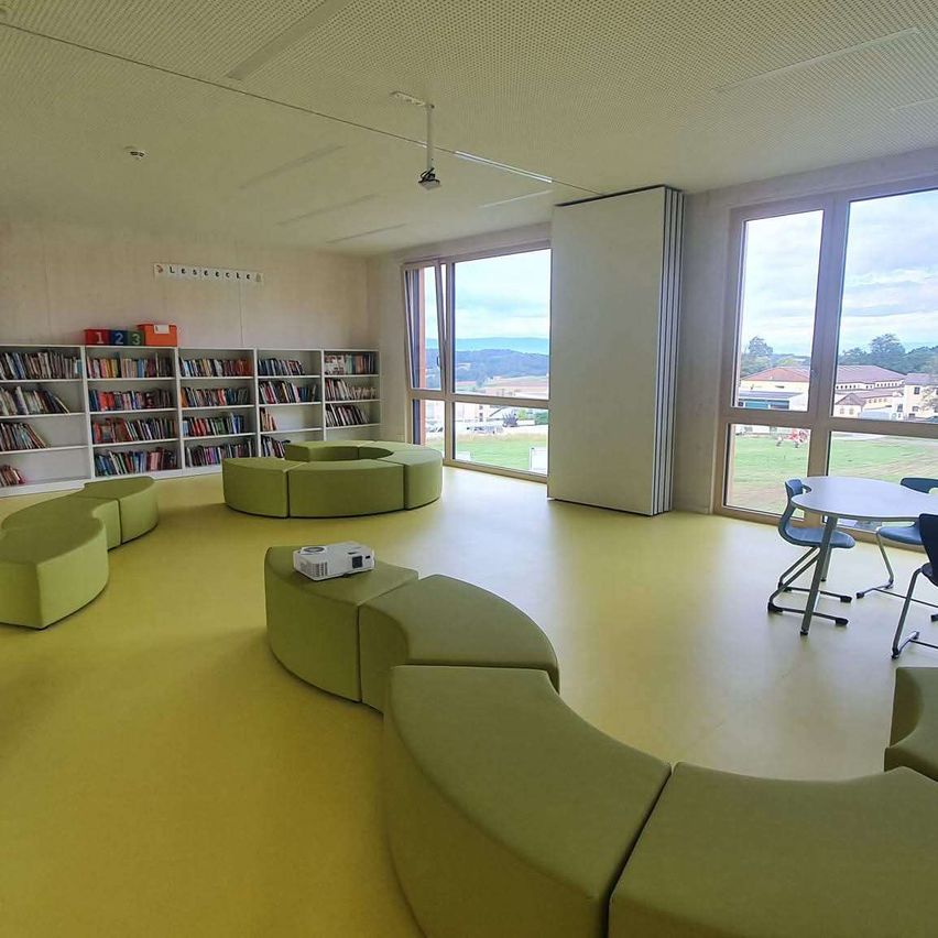 Ein heller Raum mit gelbem Boden und grüner gebogener Bestuhlung. Dahinter befinden sich mit Büchern gefüllte Bücherregale. Große Fenster bieten Blick aufs Freie.
