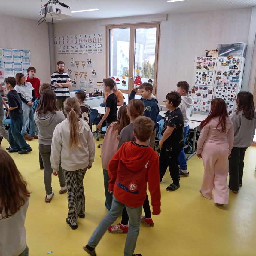Ein Klassenzimmer mit einer Gruppe von Schülern und einem Lehrer in der Mitte. Eine Projektionslampe ist an der Decke und ein Kühlschrank ist mit Aufklebern bedeckt.