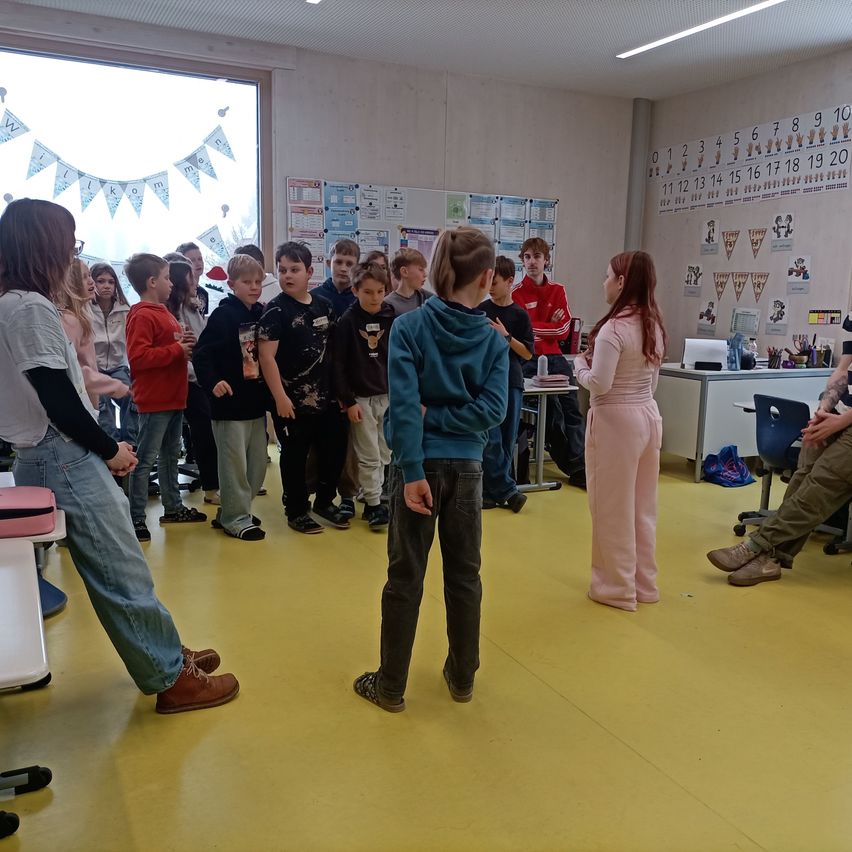 Eine Gruppe von Kindern und zwei Erwachsenen steht in einem Klassenzimmer mit gelbem Boden. Ein Erwachsener spricht zu den Kindern, während ein anderer Erwachsener auf einem Stuhl sitzt. Das Klassenzimmer hat eine Tafel mit Zahlen und eine Fahne an der Wand.