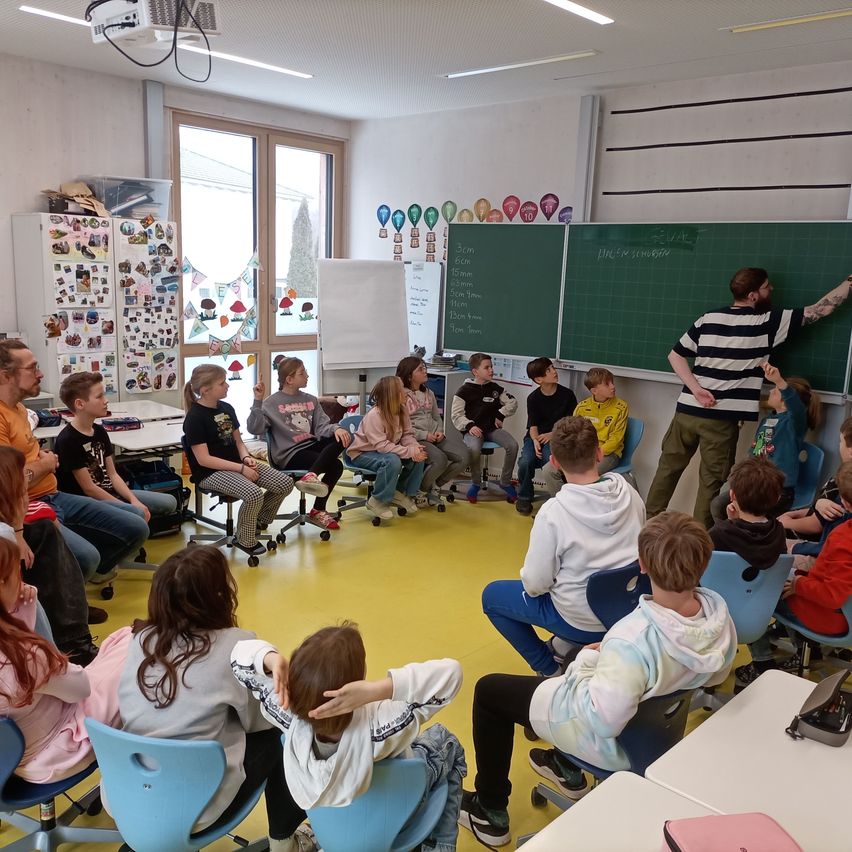 Ein Klassenzimmer mit Schülern, die auf blauen Stühlen sitzen, und einem Lehrer, der auf eine grüne Tafel schreibt. Das Klassenzimmer hat weiße Wände, gelben Boden und verschiedene Dekorationen.