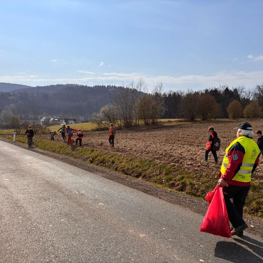 Eine Gruppe von Menschen in Warnwesten sammelt auf einem sonnigen Tag Müll in einem ländlichen Gebiet. Die Straße ist leer, und eine Person geht darauf.