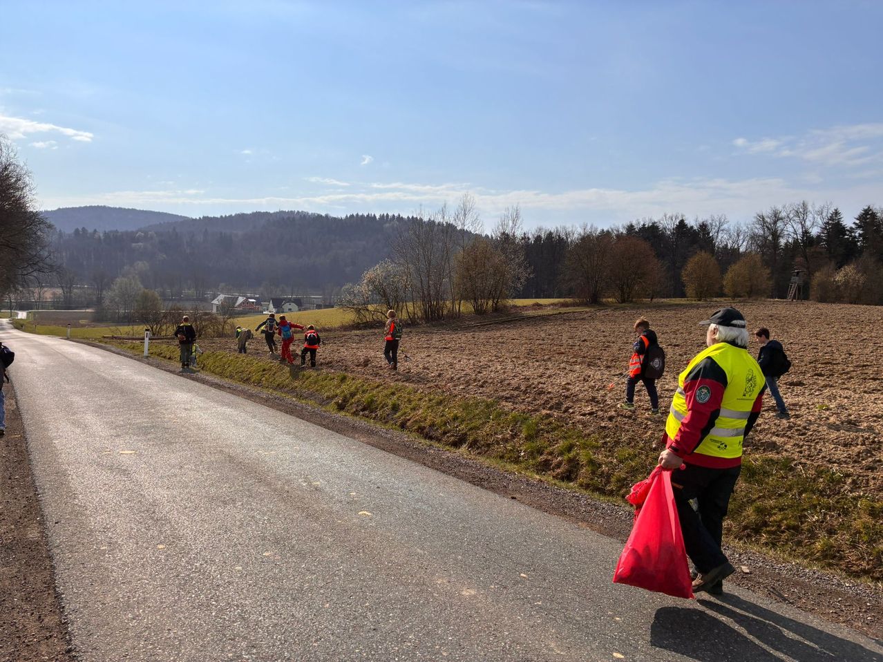 Eine Gruppe von Menschen in Warnwesten sammelt auf einem sonnigen Tag Müll in einem ländlichen Gebiet. Die Straße ist leer, und eine Person geht darauf.