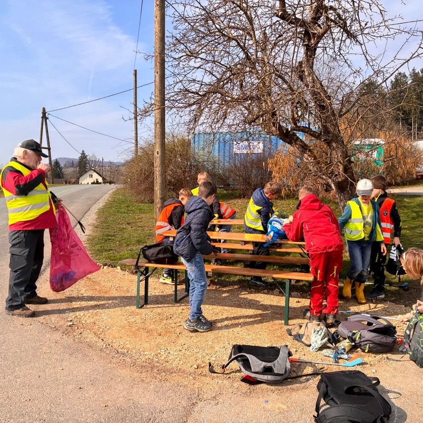 Eine Gruppe von Menschen in Sicherheitswesten und Stiefeln versammelt sich um eine Bank, einige sitzen und andere stehen. Ein Mann mit einer pinken Tasche ist in der Nähe. In der Nähe liegen Rucksäcke und Handschuhe auf dem Boden.