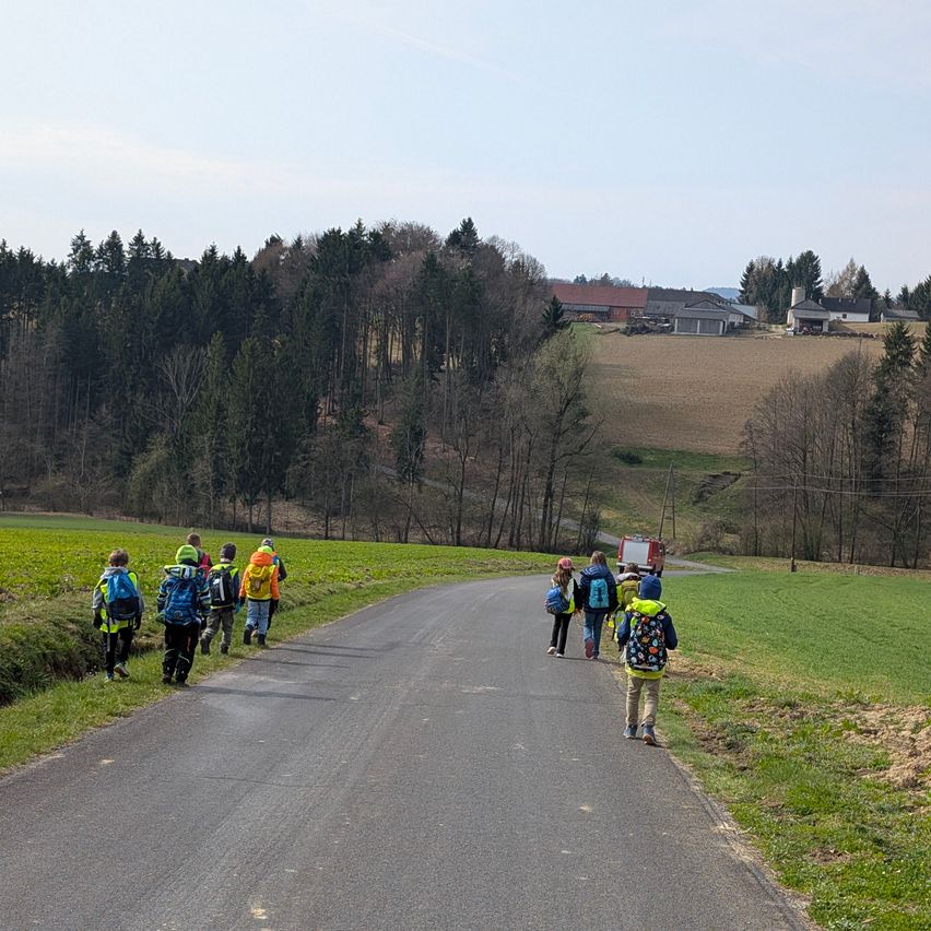 Eine Gruppe von Kindern geht mit Rucksäcken auf einer Straße, ein Feuerwehrauto ist in der Ferne sichtbar, umgeben von grünen Feldern und Bäumen.