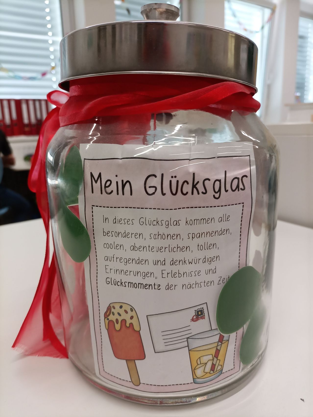 Ein Glas mit rotem Band, beschriftet 'Mein Glucksglas', gefüllt mit buntem Papier und Bildern. Der Glas hat einen Metalldeckel.