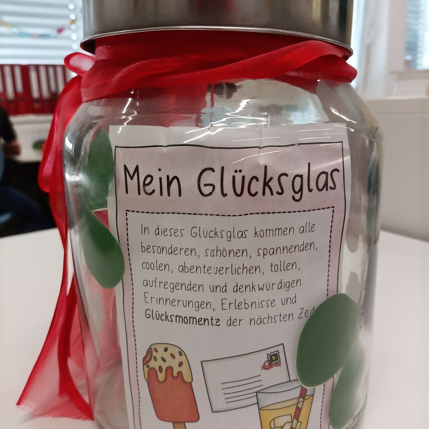 Ein Glas mit rotem Band, beschriftet 'Mein Glucksglas', gefüllt mit buntem Papier und Bildern. Der Glas hat einen Metalldeckel.