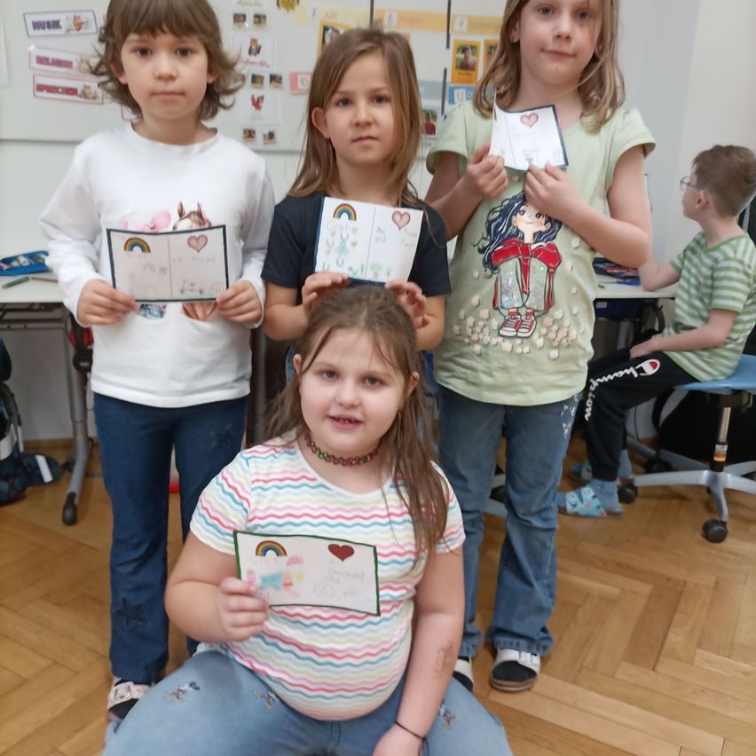 Fünf Kinder stehen in einem Klassenzimmer und halten bunte Zeichnungen. Ein Kind vorn trägt ein gestreiftes Shirt und hält eine Zeichnung mit einem Regenbogen. Hinter ihr halten drei Mädchen Zeichnungen mit Herzen. Ein weiterer Junge sitzt im Hintergrund an einem Schreibtisch.