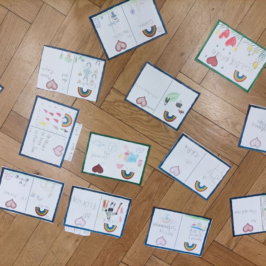 Mehrere handgezeichnete Karten mit bunten Illustrationen und Text auf einem Holzboden verteilt, einige mit Regenbögen, Herzen und Zahlen.