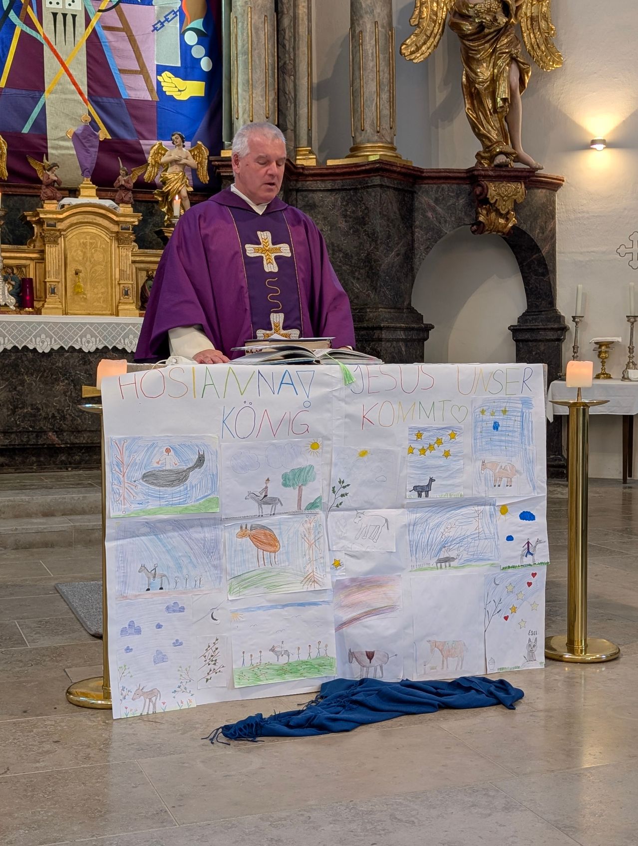 Ein Priester steht in einer Kirche und liest aus einem offenen Buch. Vor ihm steht ein großes Plakat mit Zeichnungen und der Aufschrift 'Hosanna Jesus Unser König Kommt'. Die Szene ist mit Kerzen, religiösen Symbolen und Dekorationen geschmückt.