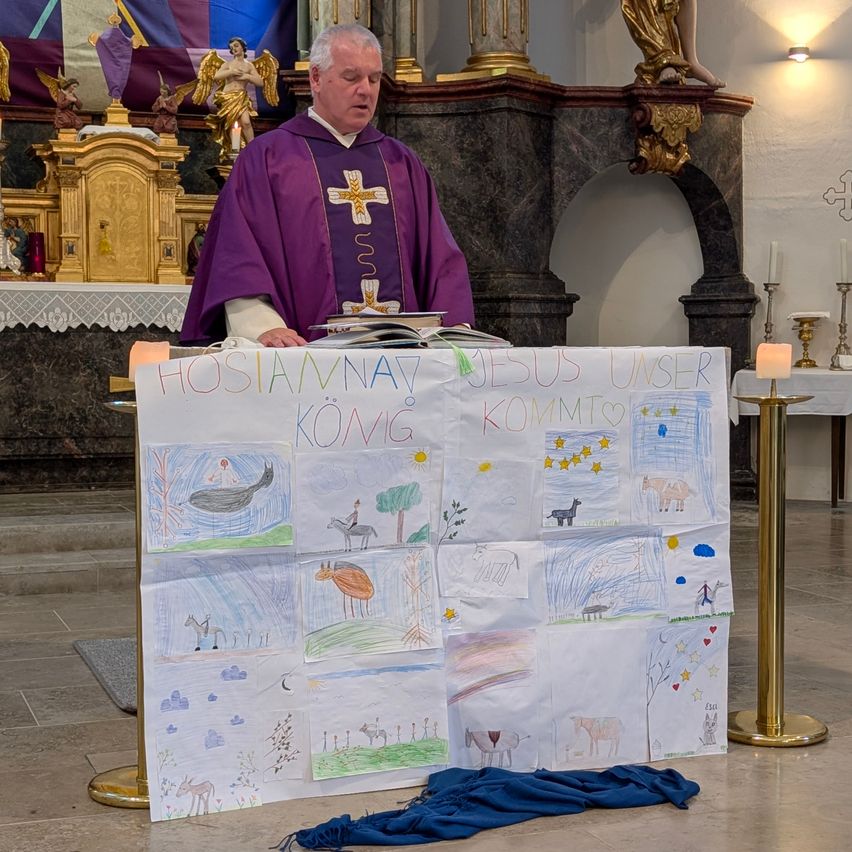 Ein Priester steht in einer Kirche und liest aus einem offenen Buch. Vor ihm steht ein großes Plakat mit Zeichnungen und der Aufschrift 'Hosanna Jesus Unser König Kommt'. Die Szene ist mit Kerzen, religiösen Symbolen und Dekorationen geschmückt.