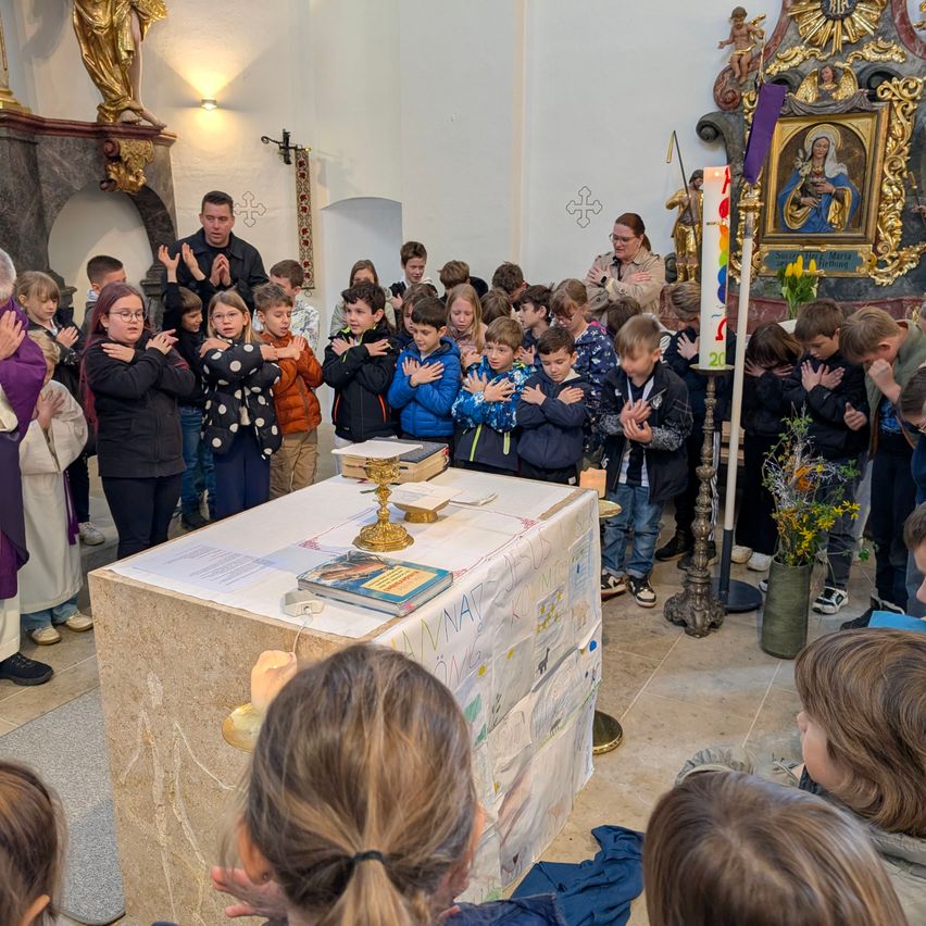 In einer Kirche stehen Kinder und Erwachsene um einen Altar, die Hände zum Gebet gefaltet. Der Altar hat einen goldenen Kerzenhalter und eine bunte Kerze. Ein Gemälde der Jungfrau Maria ist an der Wand.