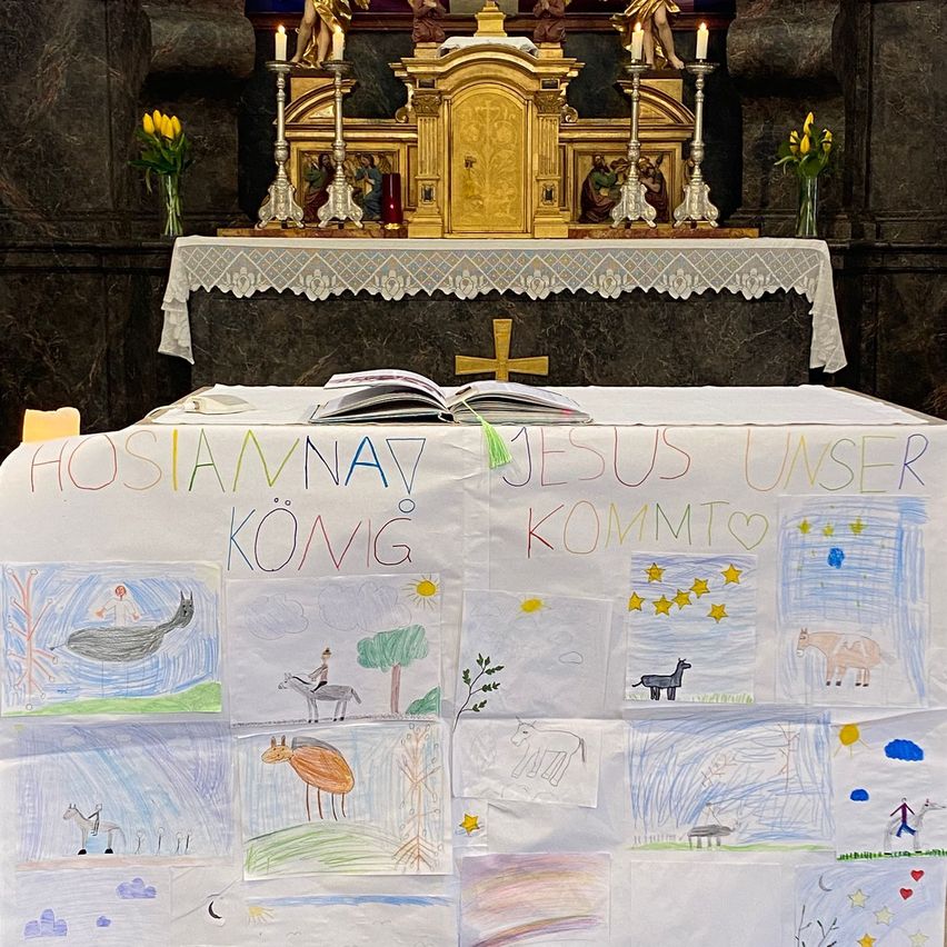 Ein Tisch mit einer weißen Tischdecke zeigt Kinderzeichnungen und die Botschaft 'Hosanna, Jesus unser König kommt.' Dahinter befindet sich ein goldener Altar mit Kerzen.