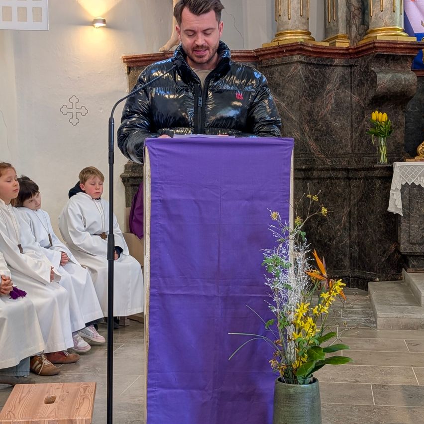 Ein Mann spricht an einer Kirchenkanzel mit einem lila Tuch. Vier Kinder in weißen Gewändern sitzen hinter ihm. Rechts steht eine Vase mit Blumen.