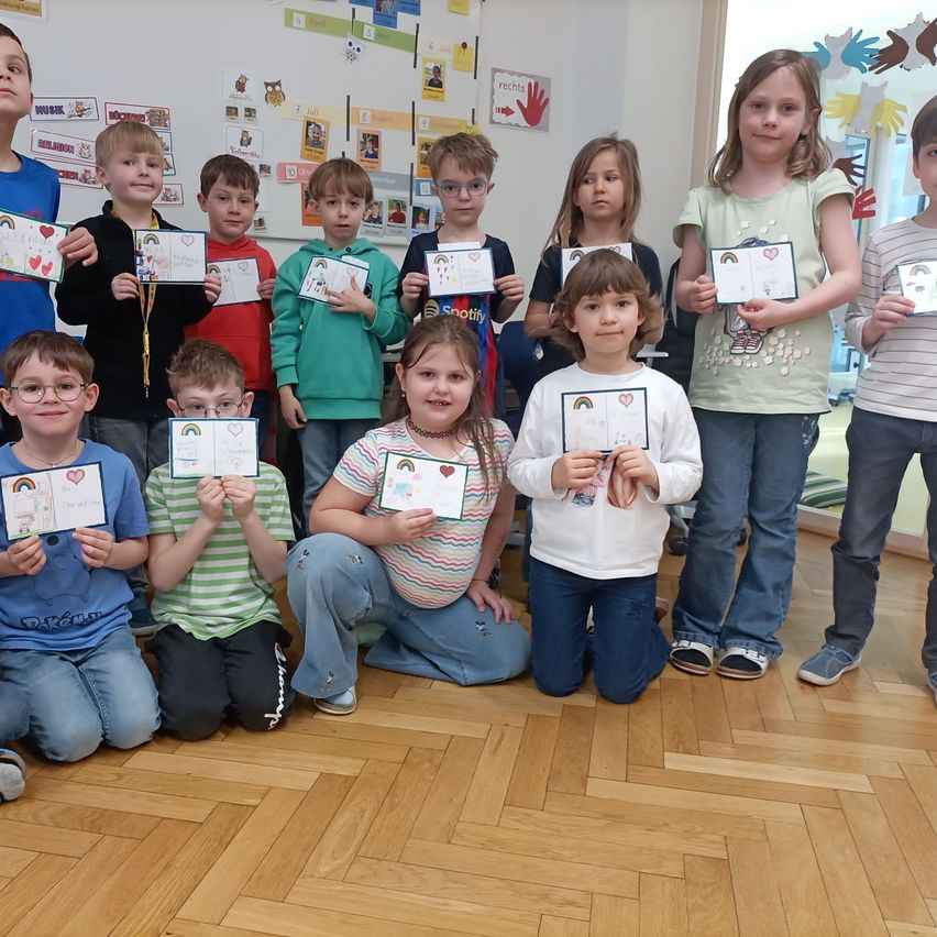 Eine Gruppe von Kindern, Jungen und Mädchen, halten Zertifikate in einem Klassenzimmer, sitzend und stehend auf einem Holzboden.