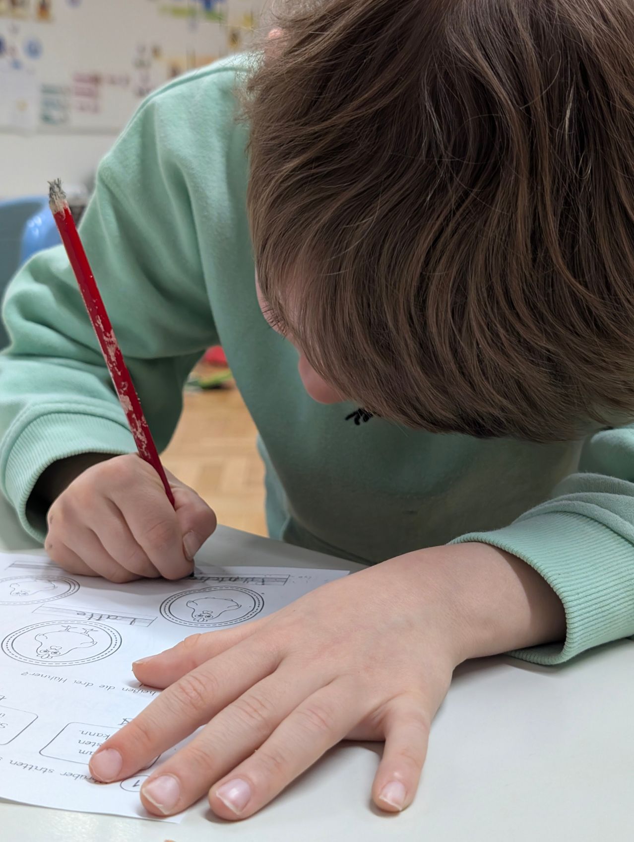 Ein Kind in einem grünen Shirt beugt sich über einen Schreibtisch und färbt ein Bild mit einem roten Stift ein.