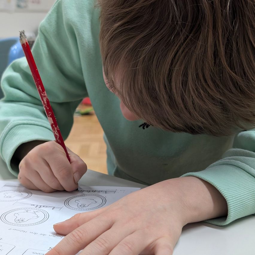 Ein Kind in einem grünen Shirt beugt sich über einen Schreibtisch und färbt ein Bild mit einem roten Stift ein.