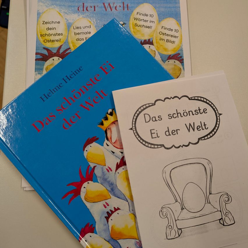 Ein blaues Buch mit dem Titel 'Das schönste Ei der Welt' liegt auf einem weißen Blatt mit einer Illustration eines Stuhls und eines Eies. Dahinter befinden sich ein weiteres Buch und ein Poster mit ähnlichen Illustrationen.