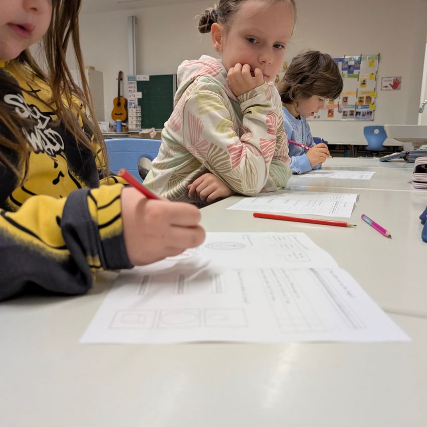Drei junge Kinder sitzen an einem Tisch und schreiben. Ein Kind hält einen Stift und scheint konzentriert zu sein. Ein anderes Kind stützt ihr Kinn auf ihre Hand, während das dritte Kind auf einem Blatt Papier schreibt. Der Tisch ist mit Papieren und Stiften übersät. Im Hintergrund sind eine Gitarre und eine Tafel zu sehen.