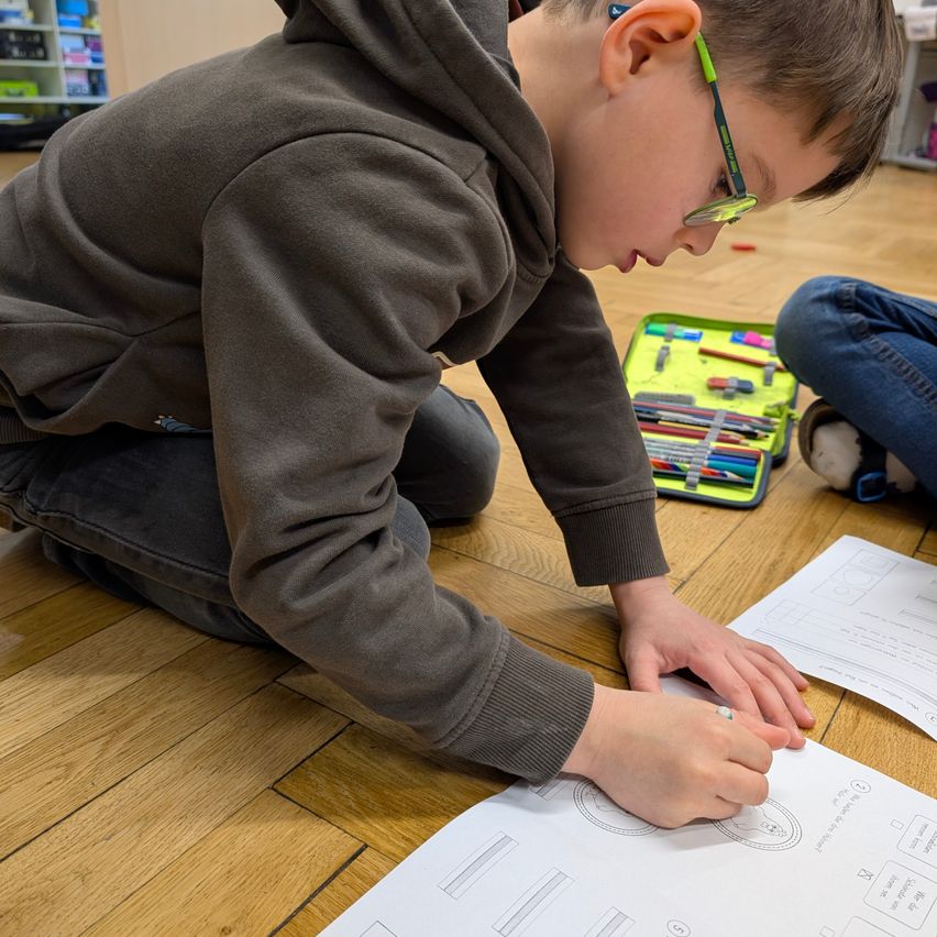 Ein Junge mit einer Brille und einem Kapuzenpullover kniet auf einem Holzboden und arbeitet an einem Papier. Ein Stiftetui mit verschiedenen farbigen Stiften steht neben ihm. Eine andere Person sitzt in der Nähe und beschäftigt sich ebenfalls mit Papieren.