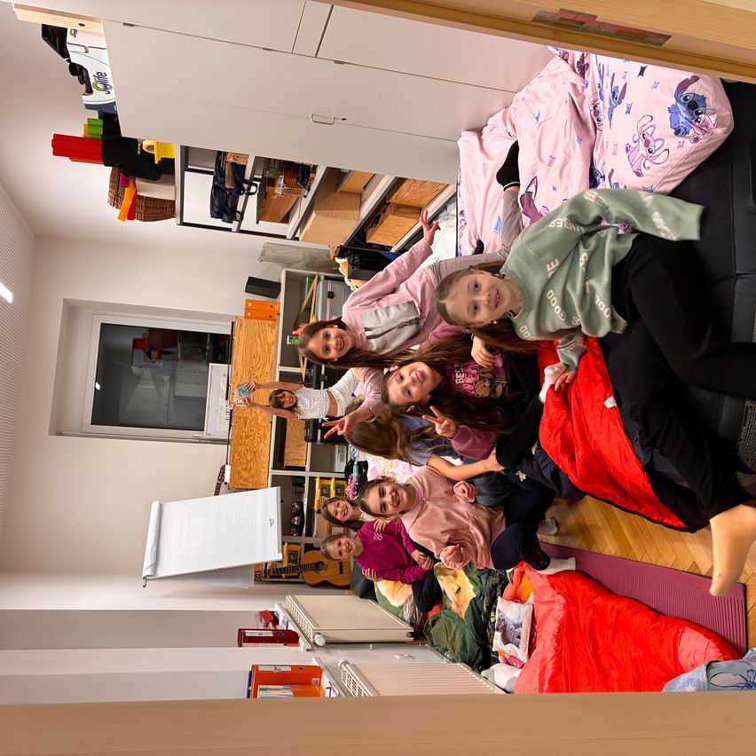 Eine Gruppe von Kindern liegt auf dem Boden in einem Zimmer, möglicherweise bei einem Schlafübernachtungsfest. Sie sind von verschiedenen Gegenständen und Utensilien umgeben.