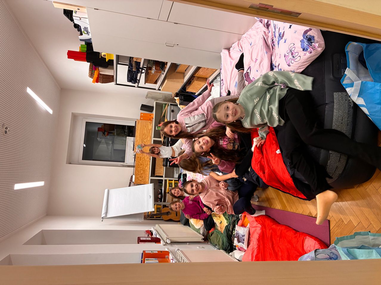 Eine Gruppe von Kindern liegt auf dem Boden in einem Zimmer, möglicherweise bei einem Schlafübernachtungsfest. Sie sind von verschiedenen Gegenständen und Utensilien umgeben.