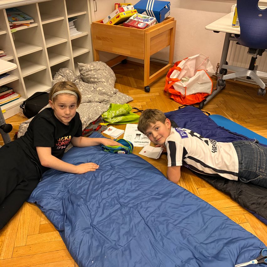 Zwei Kinder liegen auf Schlafsäcken auf einem Holzboden. Sie haben Bücher, Taschen und einen kleinen Tisch im Hintergrund. Der Junge lächelt, während das Mädchen ihn ansieht.