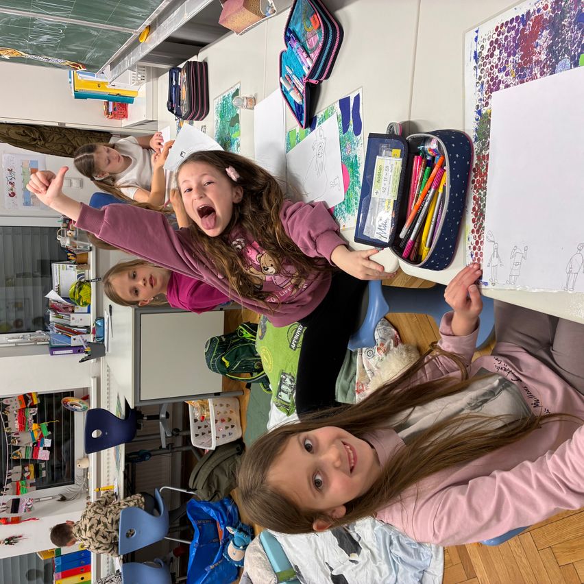 Drei junge Mädchen lächeln und posieren für die Kamera in einem Klassenzimmer. Sie sitzen an einem Tisch mit Zeichnungen, Stiften und anderen Schulmaterialien.